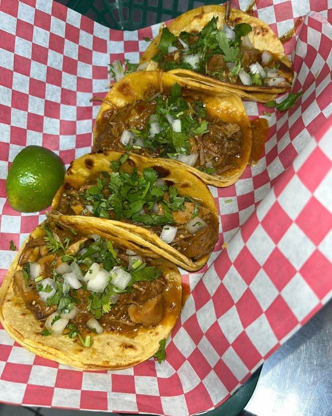 Four tacos topped with cilantro and onions in a red-and-white checkered basket with a lime wedge.