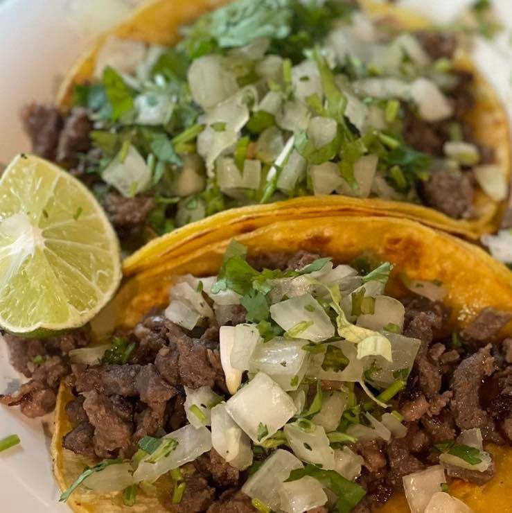 Two tacos on corn tortillas topped with chopped onion, cilantro, and a lime wedge.