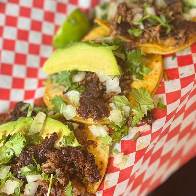 Three street tacos topped with meat, cilantro, onions, and avocado slices, served in a red-checkered paper tray.