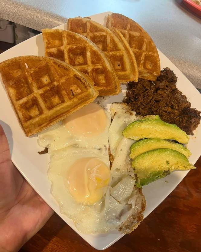 A white plate holding three waffle quarters, two sunny-side-up eggs, shredded meat, and three slices of avocado.