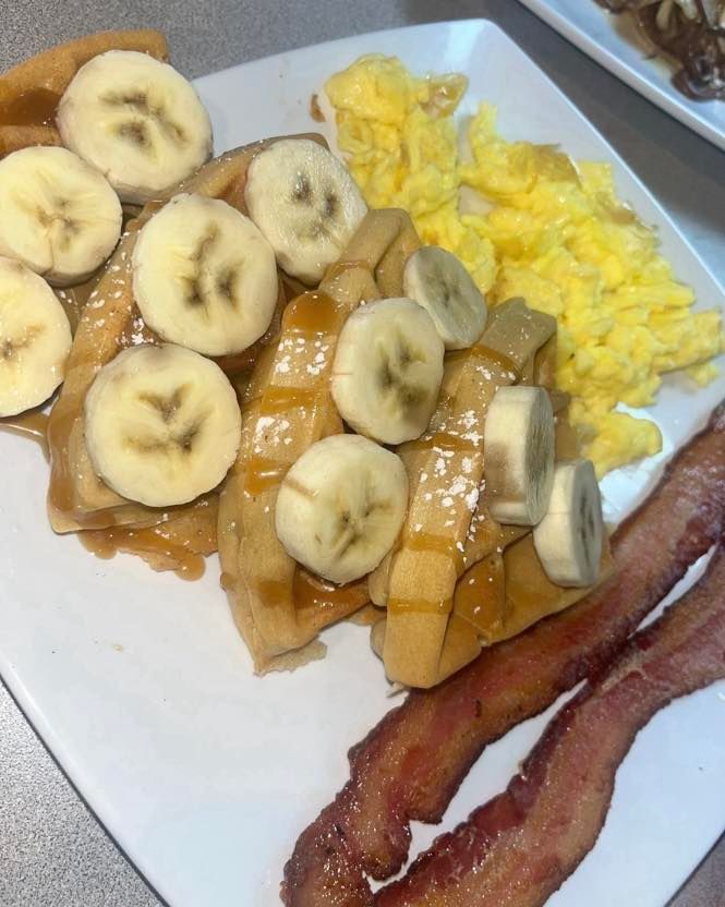 A breakfast plate featuring waffles topped with sliced bananas and syrup, served with scrambled eggs and two bacon strips.
