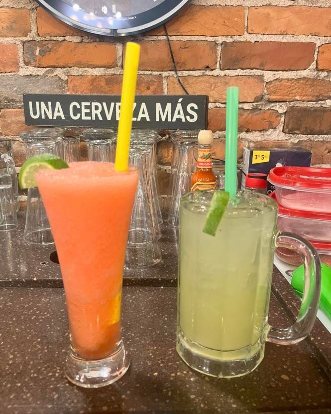 A frozen pink drink with a lime wedge and yellow straw next to a light yellow drink in a glass mug with a green straw.