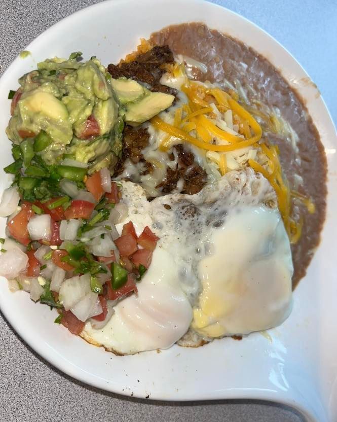 A white plate containing eggs, refried beans, guacamole, and a pico de gallo salsa mix topped with melted shredded cheese.