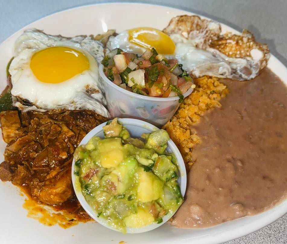 A plate featuring two fried eggs, a stewed meat dish, refried beans, Mexican rice, a side of pico de gallo, and guacamole.