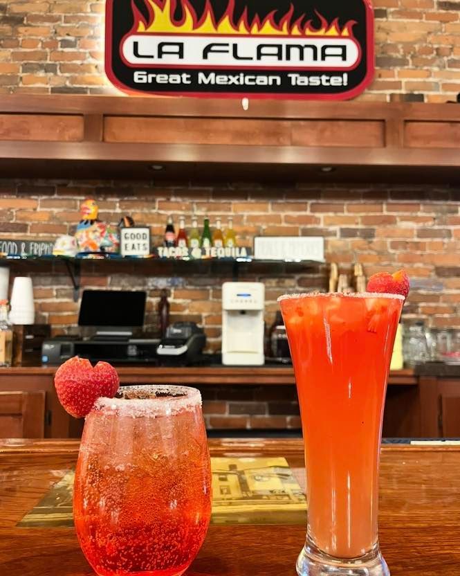 Two cocktails garnished with strawberries sit on a wooden bar in front of a brick wall sign for La Flama Mexican restaurant.