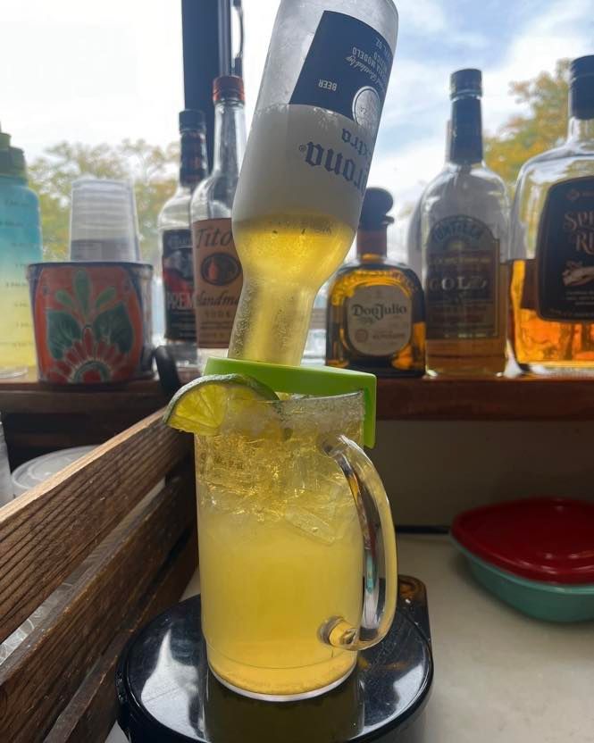 A Corona beer bottle upside down in a cocktail mug with ice, lime, and a salted rim on a bar counter with liquor bottles.