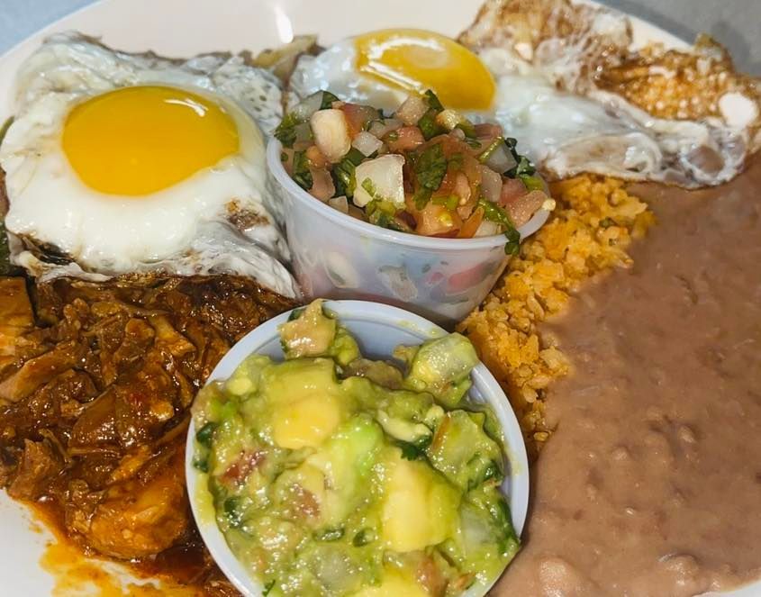 A breakfast plate with two fried eggs, stewed meat, Mexican rice, refried beans, salsa, and guacamole in small cups.