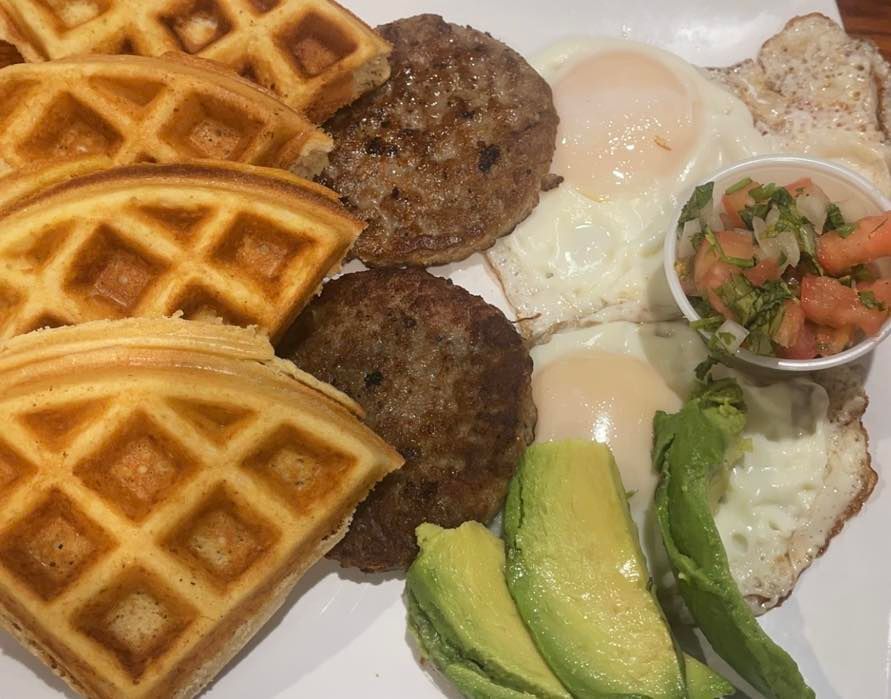 A breakfast plate with waffle quarters, two sausage patties, fried eggs, avocado slices, and a side of pico de gallo.