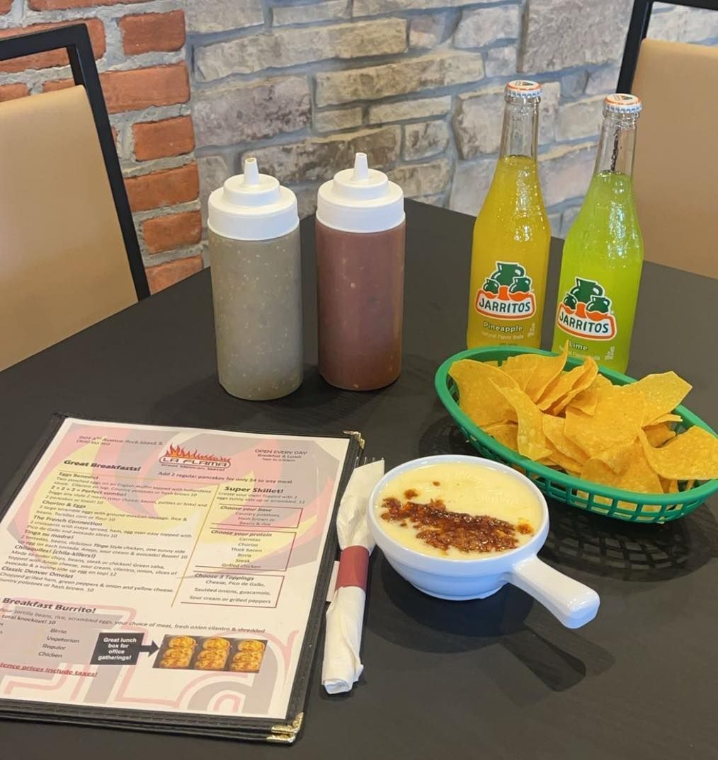 A restaurant table with a menu, chips, queso, two squeeze bottles, and two yellow Jarritos sodas against a brick wall.