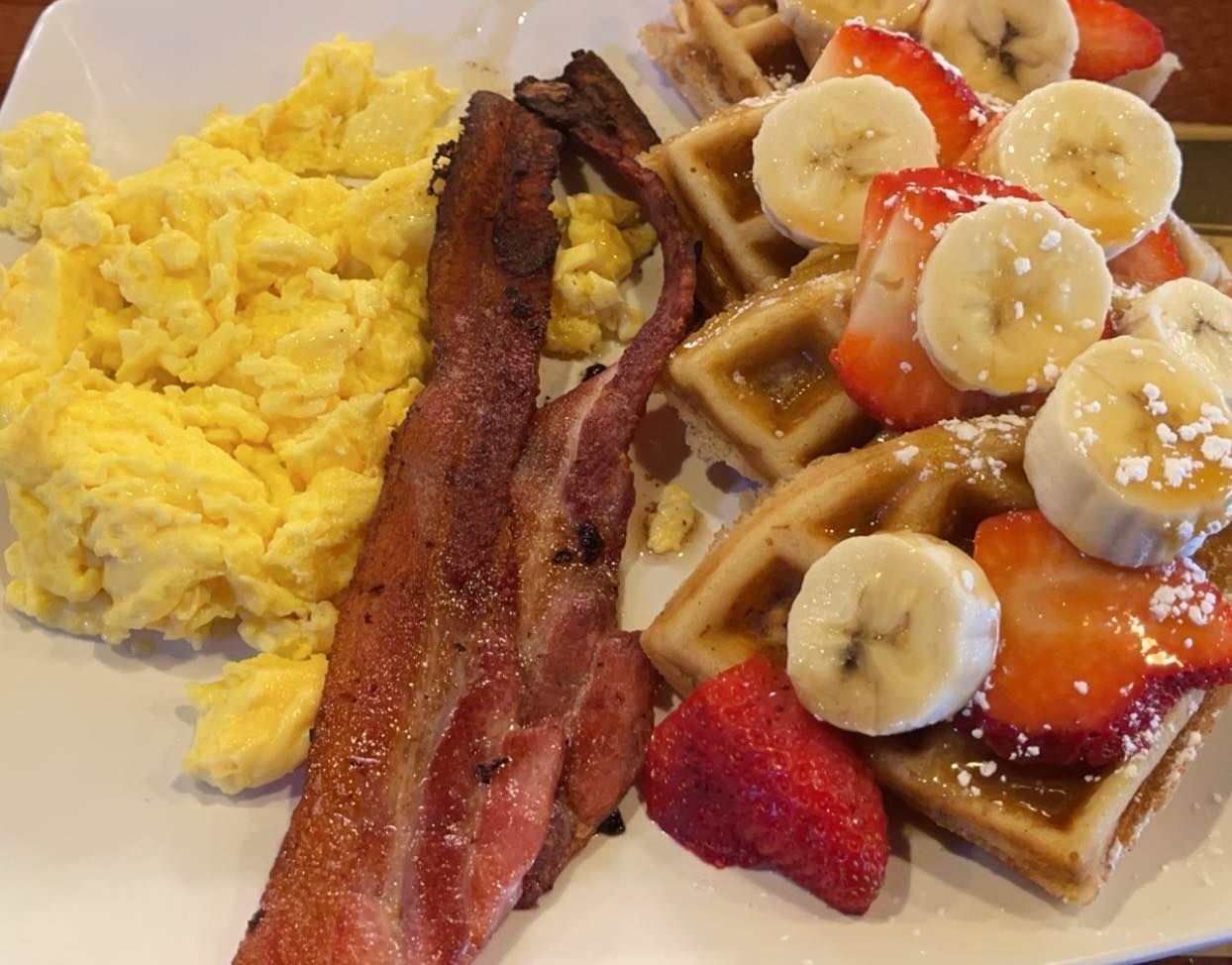 A plate of breakfast featuring scrambled eggs, two strips of bacon, and waffles topped with banana slices and strawberries.