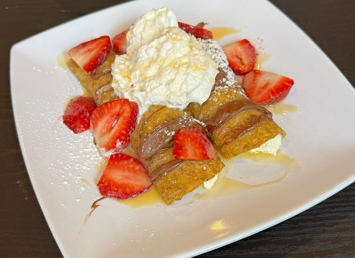 Two rolled crepes topped with a dollop of whipped cream, sliced strawberries, a chocolate drizzle, and powdered sugar.