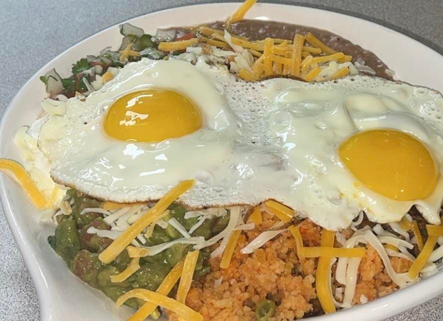 Two sunny-side-up eggs served over refried beans, rice, guacamole, and shredded cheddar cheese on a white plate.