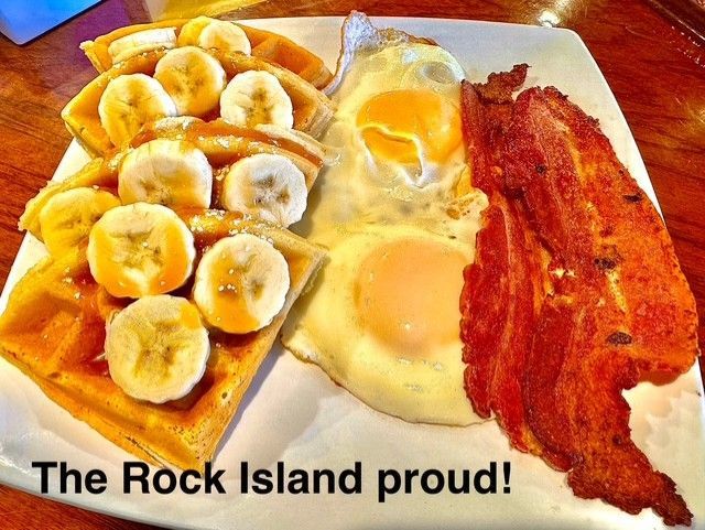 A plate of waffles topped with banana slices, served with two fried eggs and crispy bacon, labeled "The Rock Island proud!"