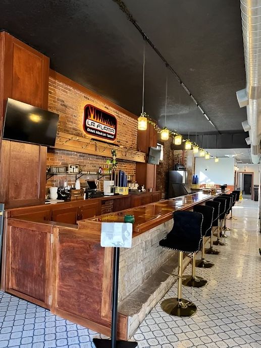 A wooden bar with stone front and black chairs, accented by glowing pendant lights and a logo on an exposed brick wall.