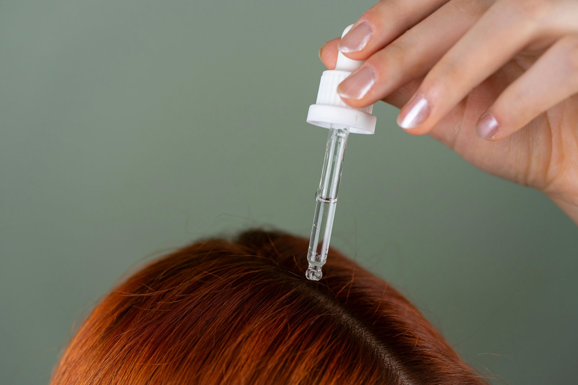 A hand with a dropper applies liquid to the scalp of red hair.