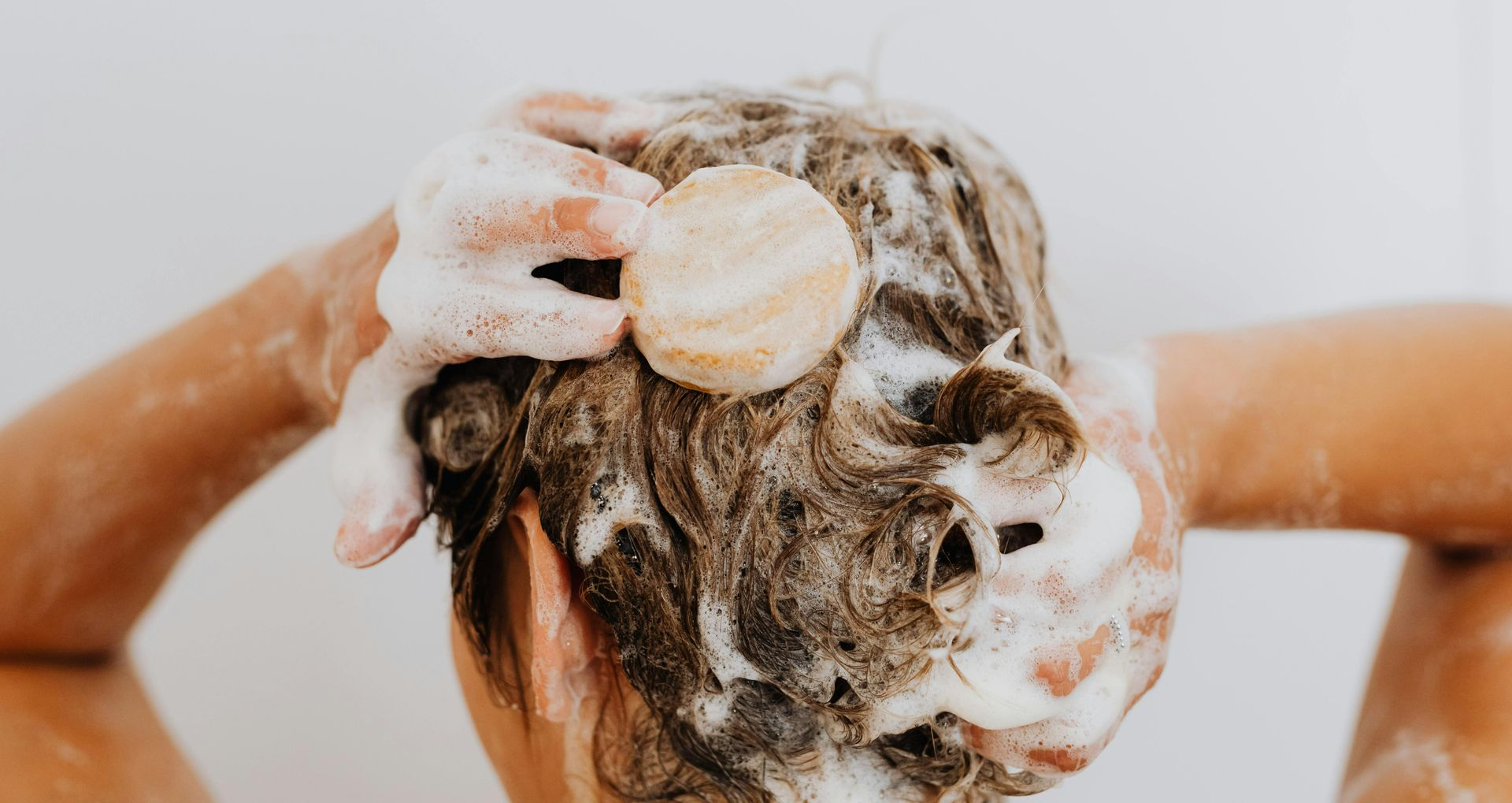 Person washing hair with a round shampoo bar, lathering foam, against a white backdrop.