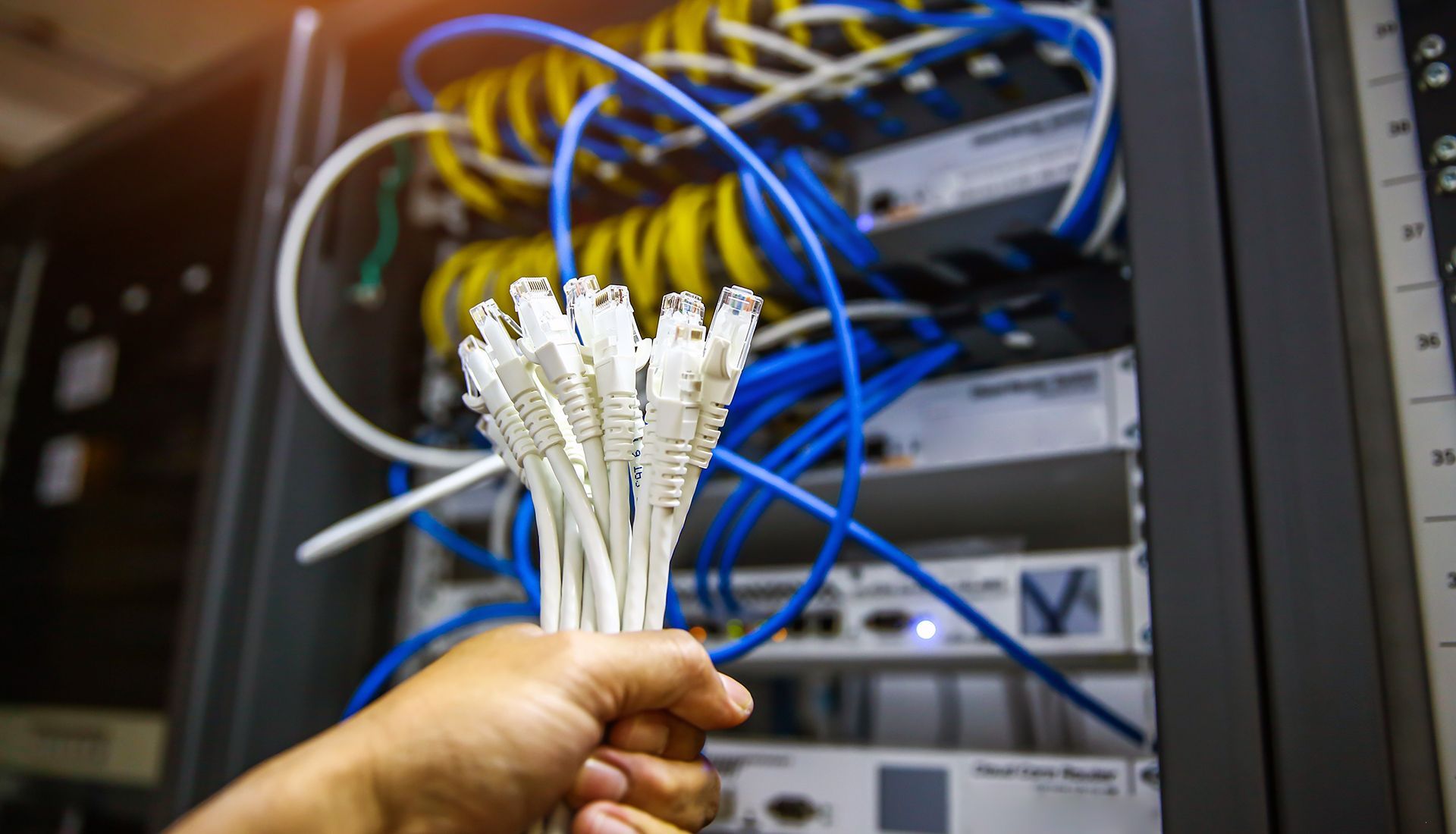 Hand holding a bundle of white Ethernet cables in front of a server rack with colorful cables.