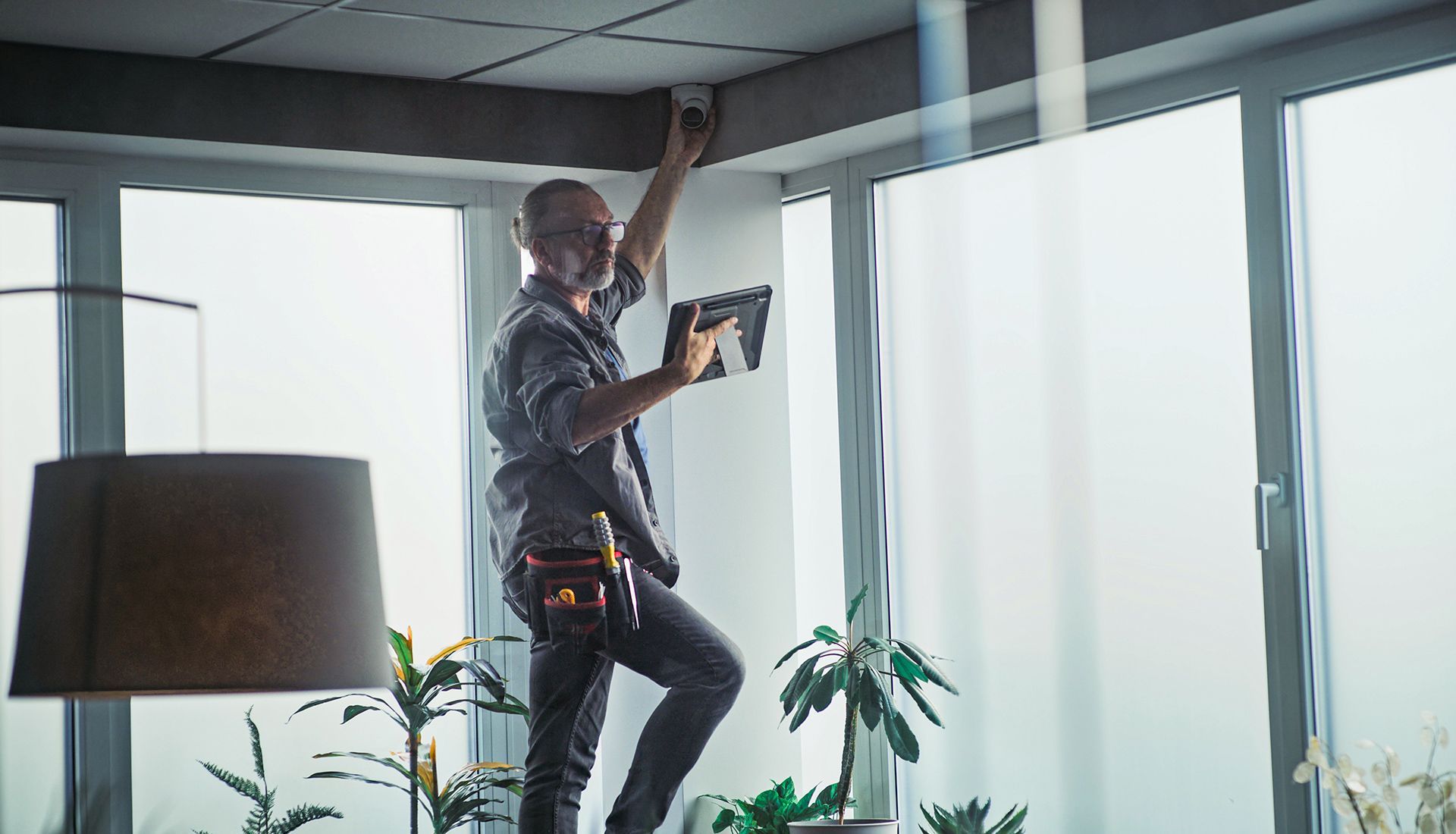 Man on ladder, installing something on ceiling near window. Holds tablet, wearing tool belt.