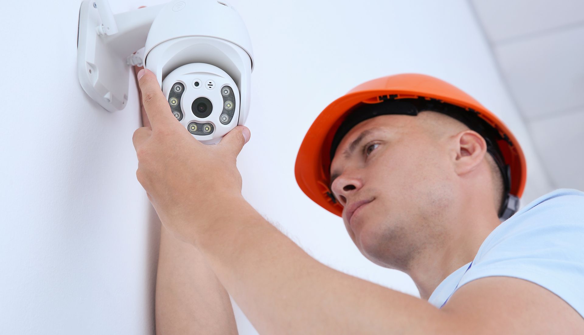 Man in orange hard hat installing a white security camera on a white wall.