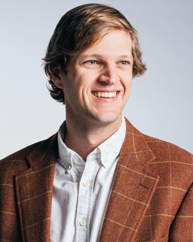 A man wearing a brown jacket and a white shirt is smiling