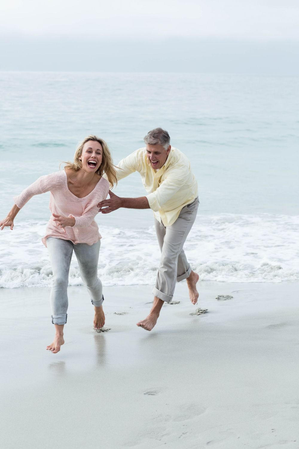 Couple running and laughing on a beach, woman in pink, man in yellow, ocean in background.