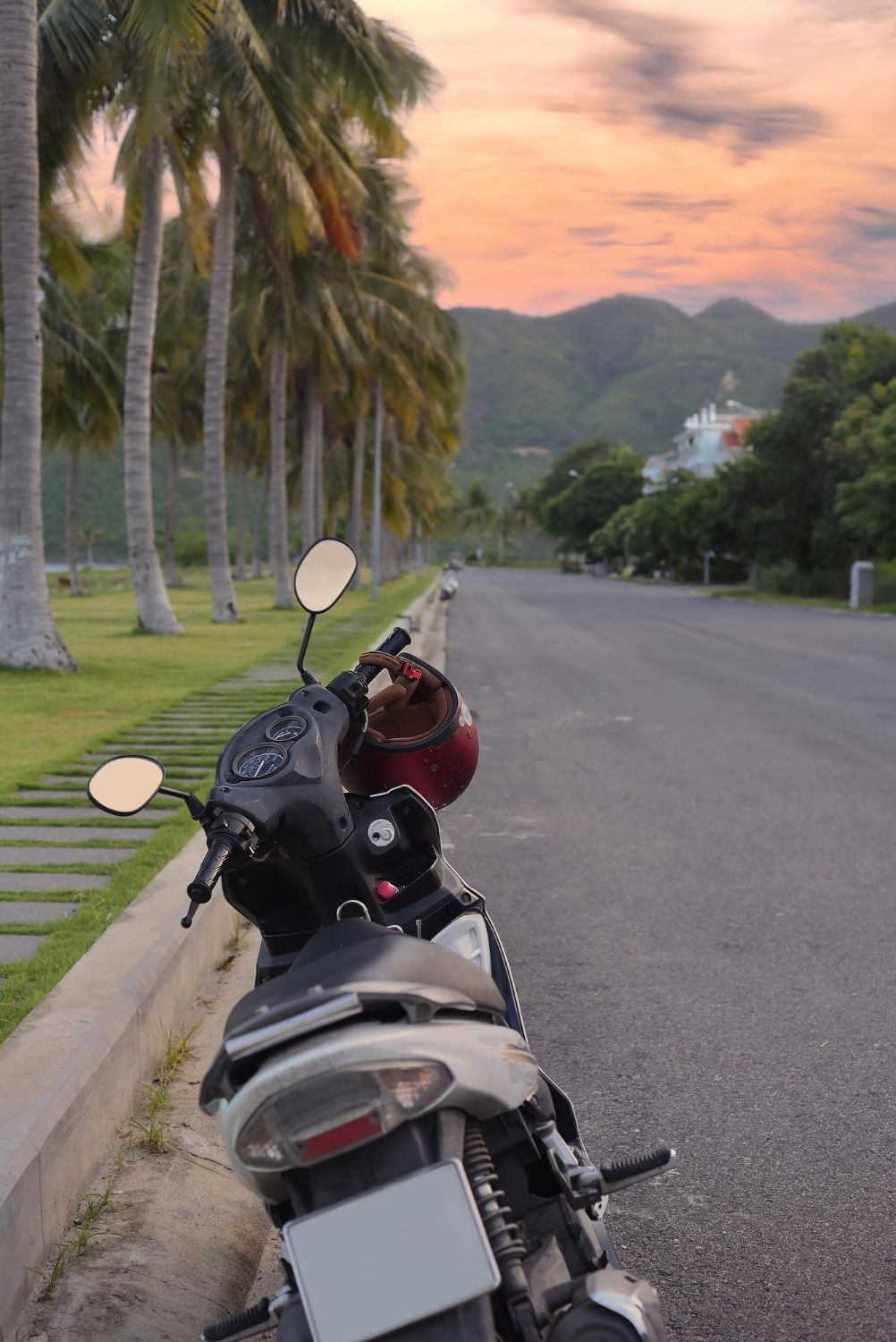 Motor scooter parked on road lined with palm trees, mountains in background, sunset sky.