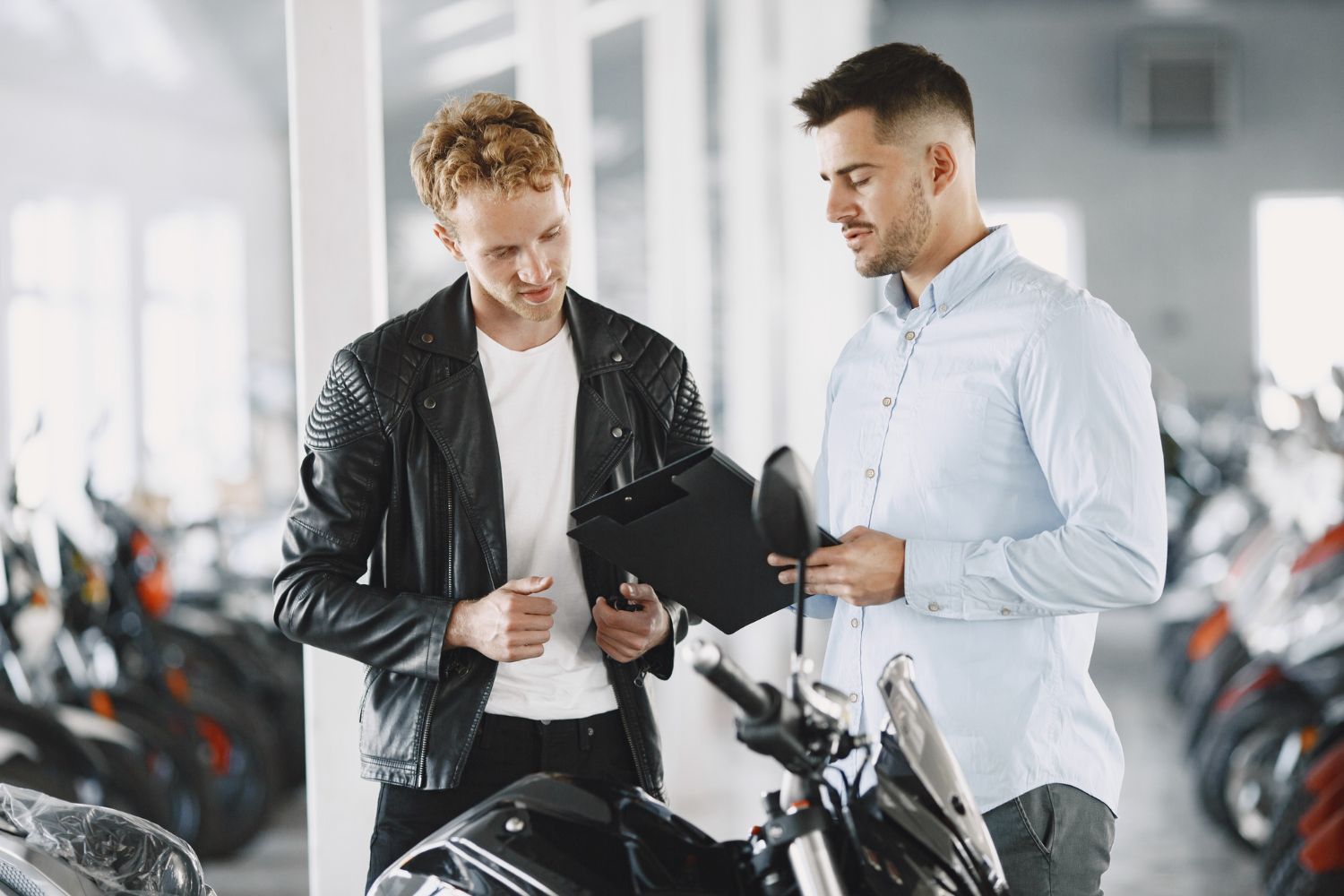 Man in leather jacket reviews motorcycle with salesperson. Showroom setting.
