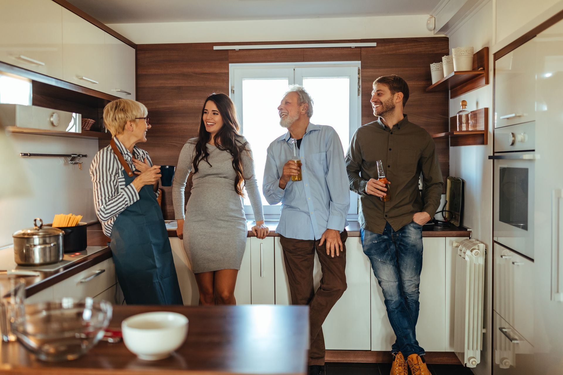 Four people in a kitchen talking, some holding drinks. Light pours in from a window.