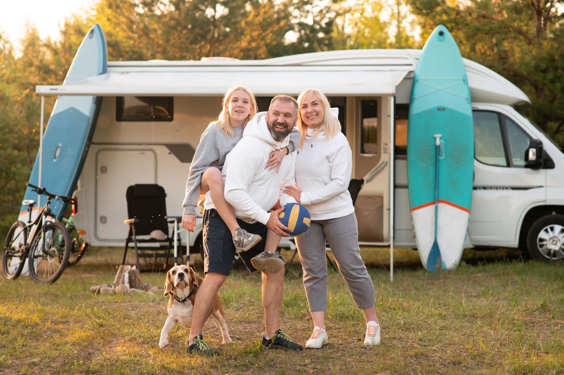 Family with dog, holding volleyball, posing in front of a camper with surfboards.