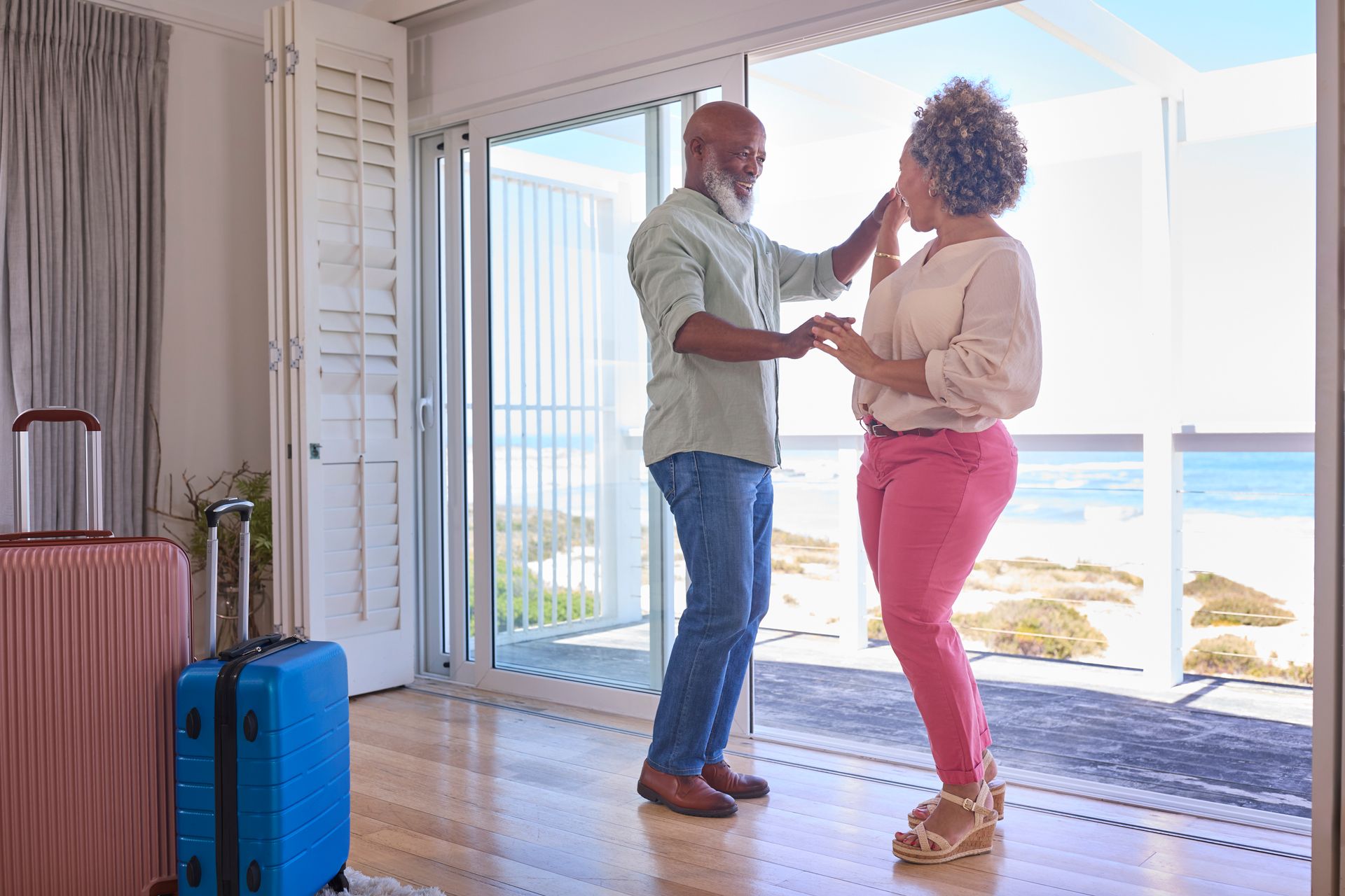 Couple dances by open patio doors overlooking the ocean, suitcases nearby.