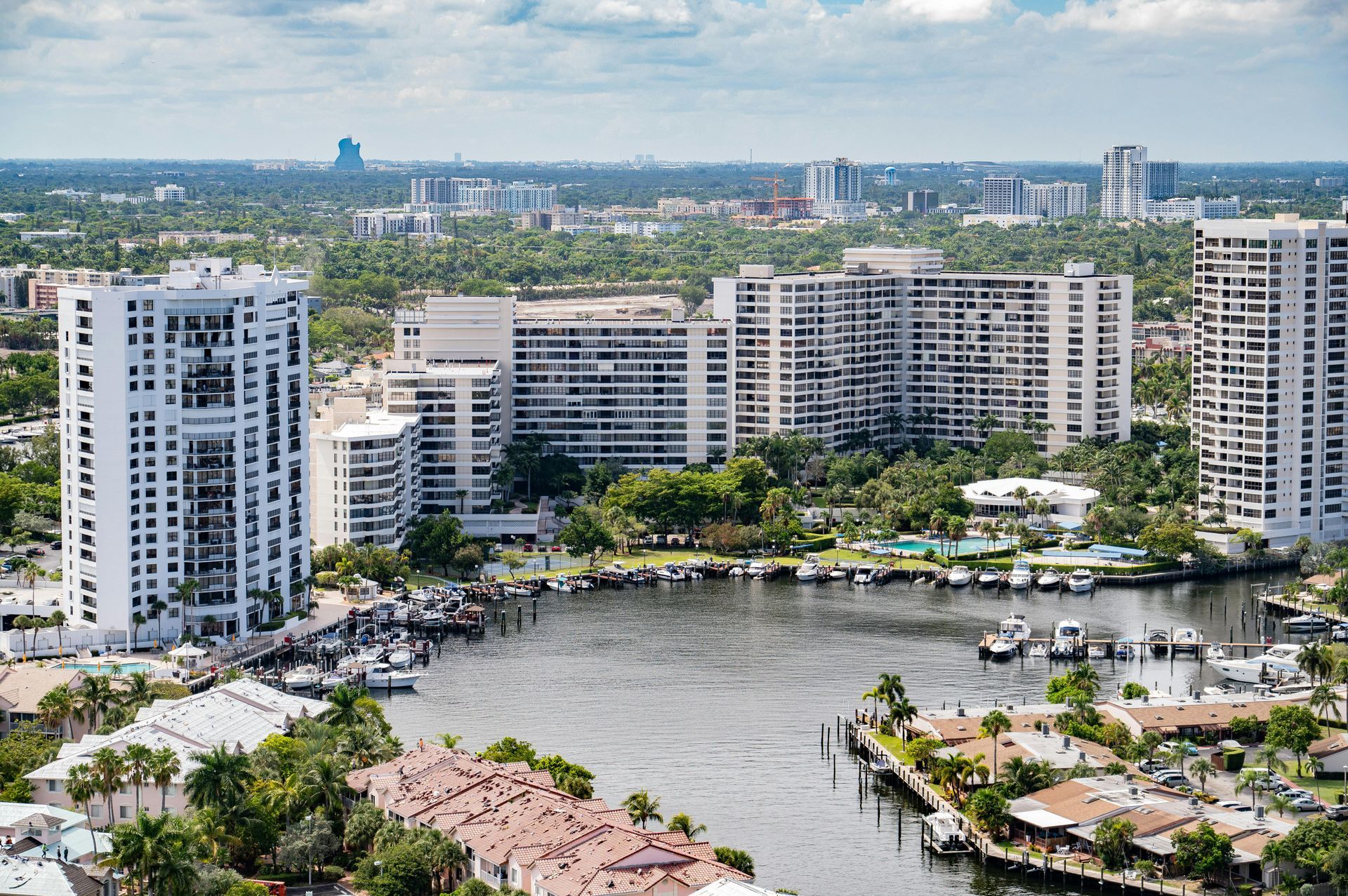 Coastal cityscape with white high-rise buildings, boats in a waterway, and lush green trees under a cloudy sky.