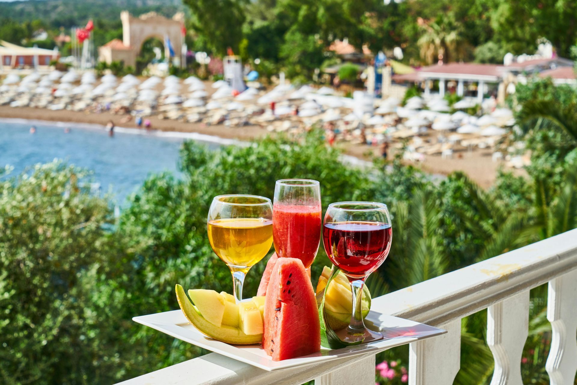 Three glasses of juice and fruit on a tray overlooking a beach.