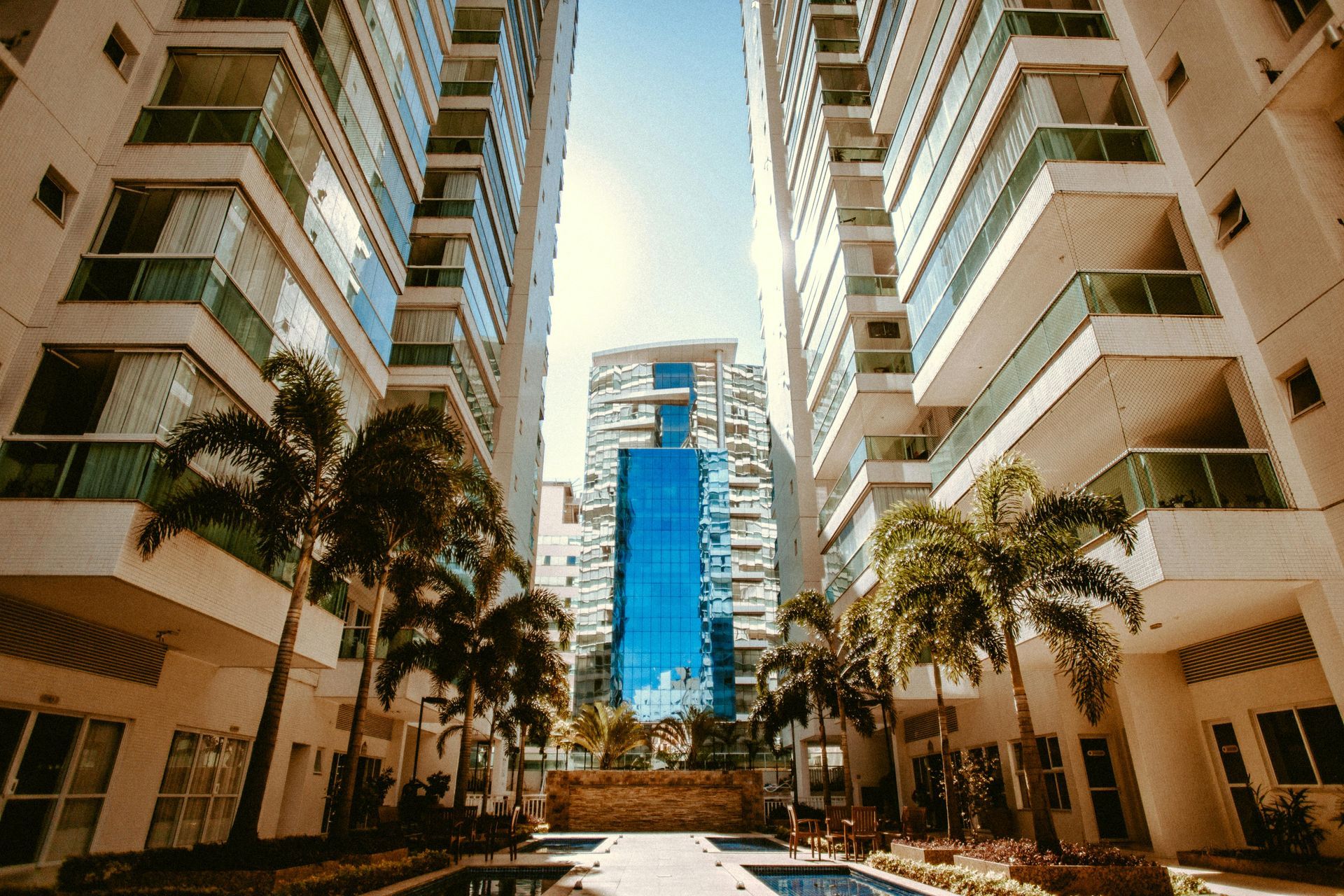 Tall buildings with balconies flank a view of a modern skyscraper; palms, sky and courtyard.