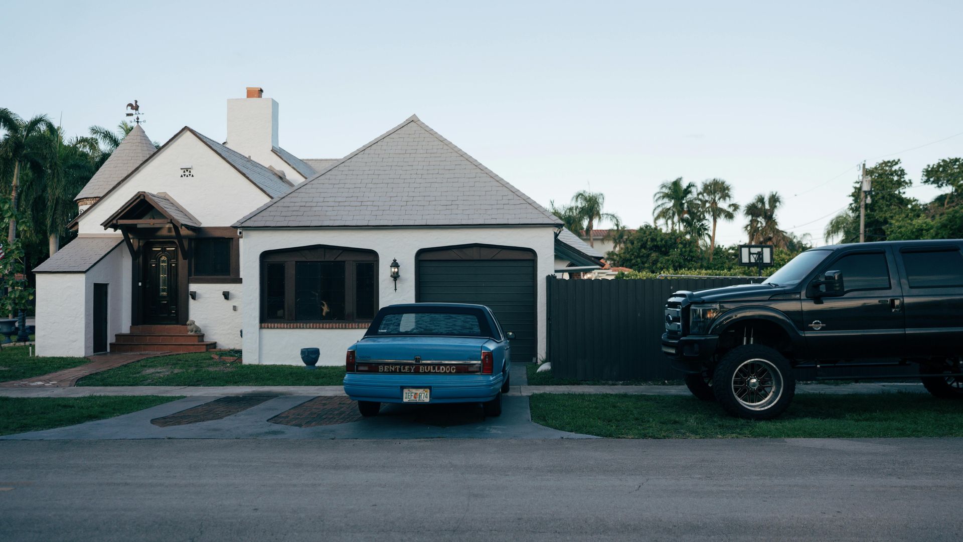 White house with blue car in driveway, black truck parked on lawn, green trees in background.