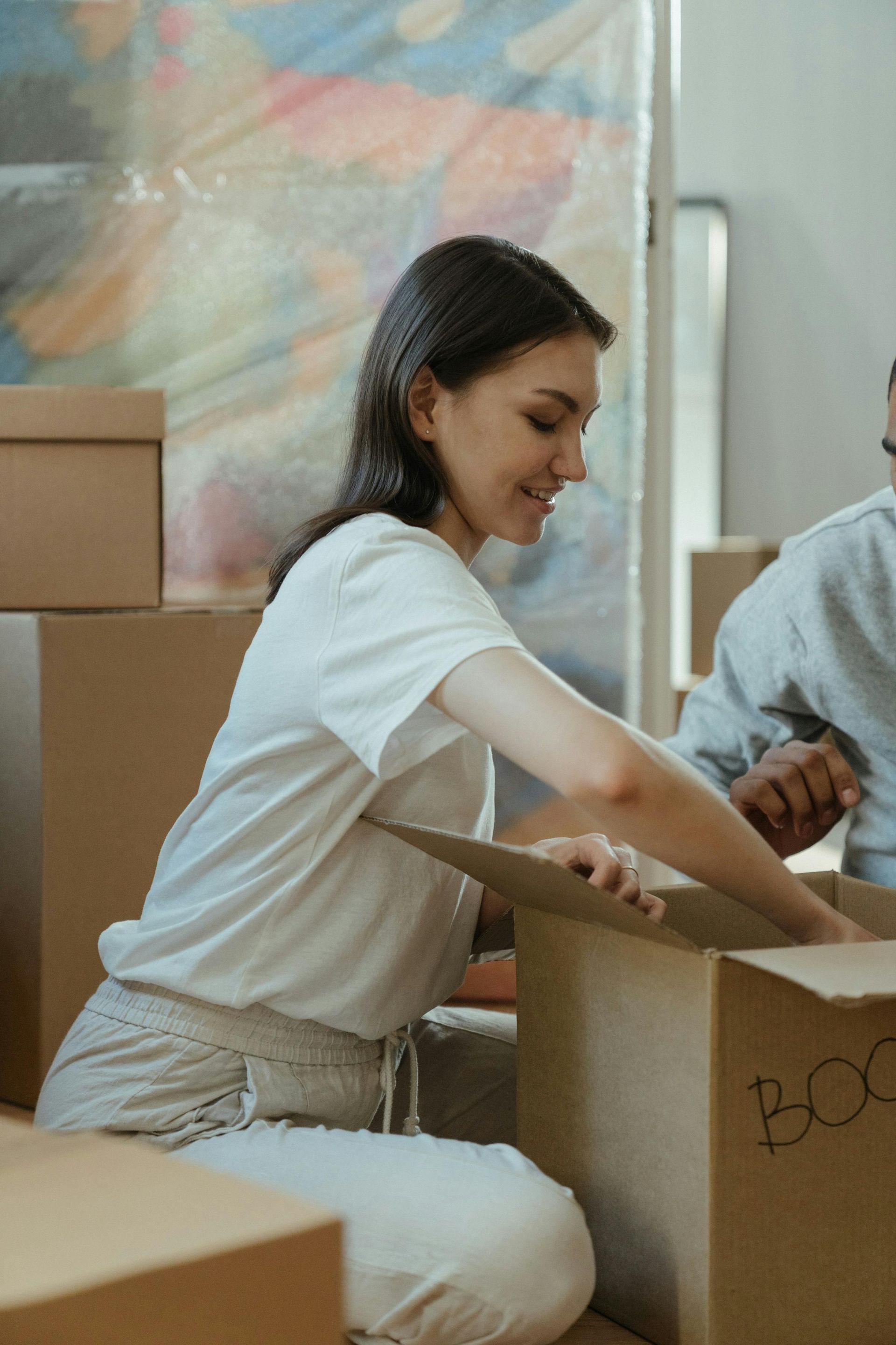 Woman packing a cardboard box, smiling. Boxes and another person nearby, in a room, light setting.