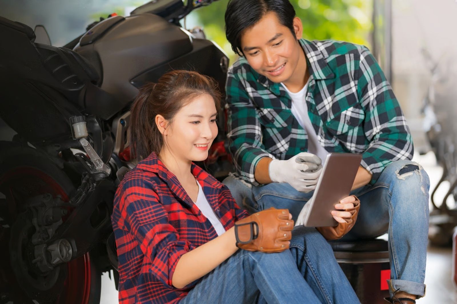 Woman and man in a garage looking at a tablet near a motorcycle. Both wearing flannels, smiling.
