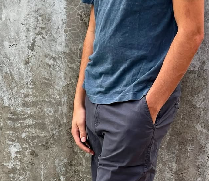 Un hombre con una camisa azul y pantalones grises está de pie con las manos en los bolsillos.