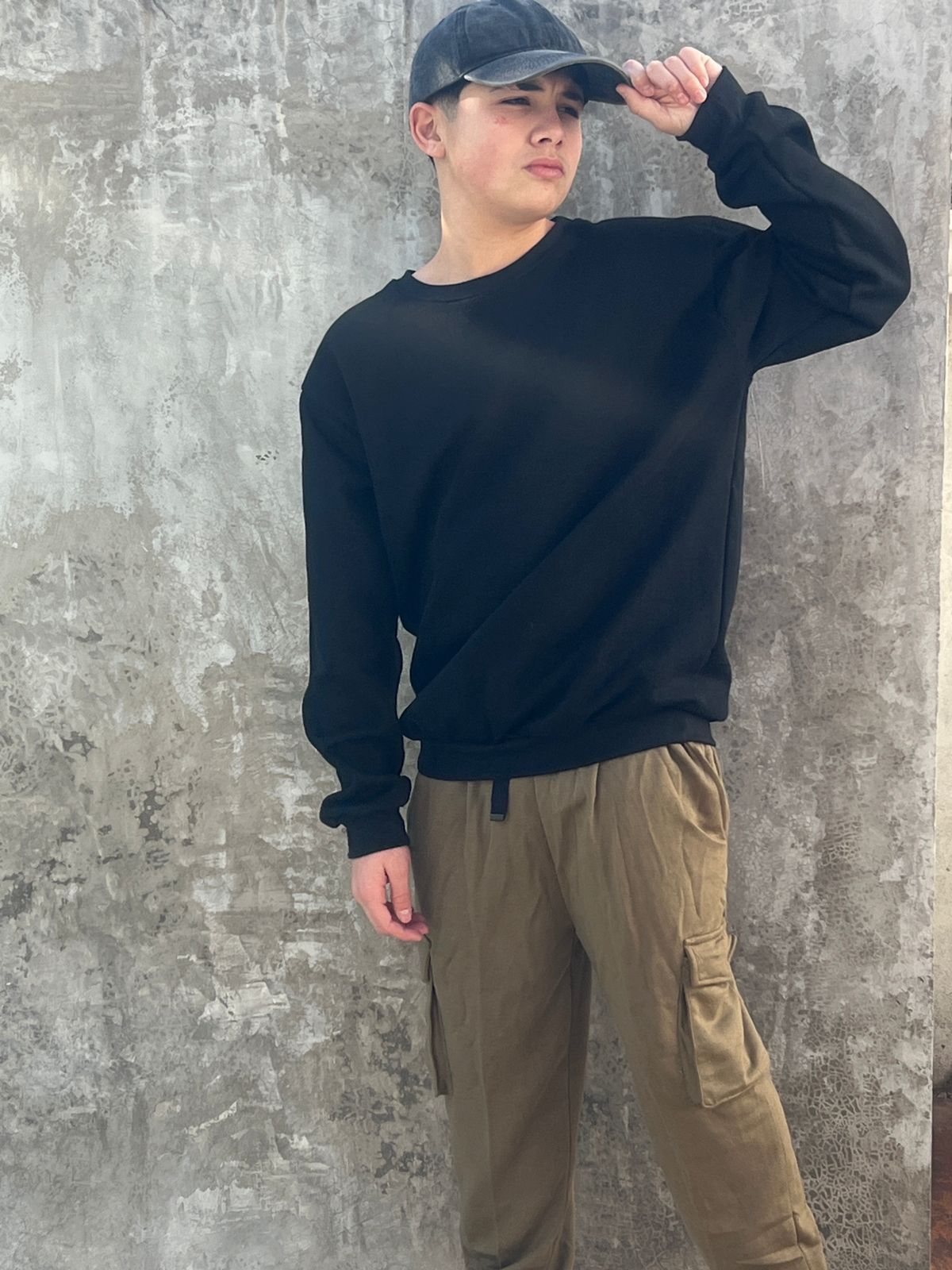 Un joven vestido con una sudadera negra y un sombrero está parado frente a un muro de hormigón.