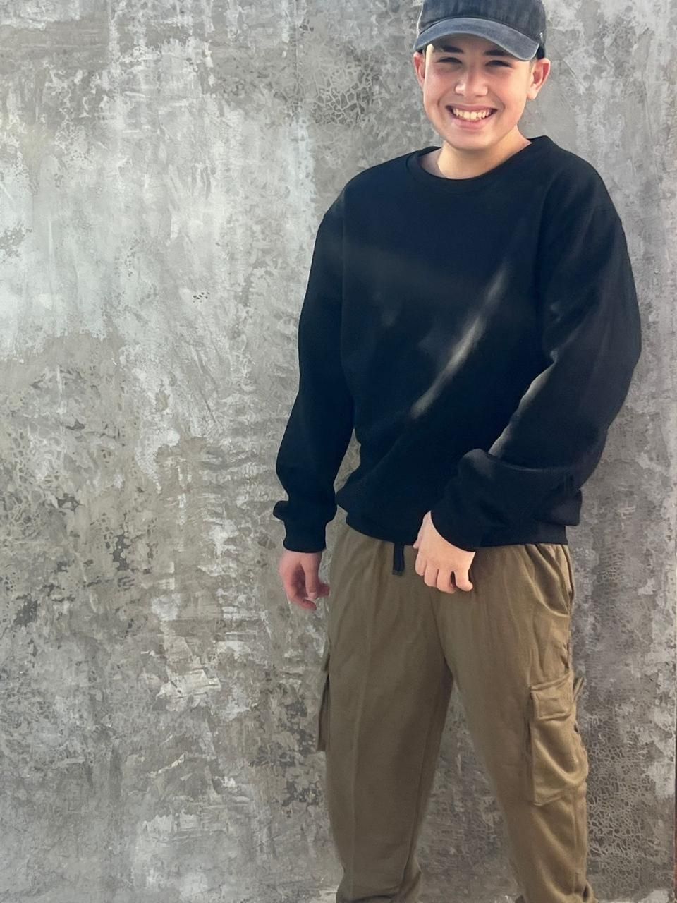 Un joven vestido con una sudadera negra y un sombrero está parado frente a un muro de hormigón.