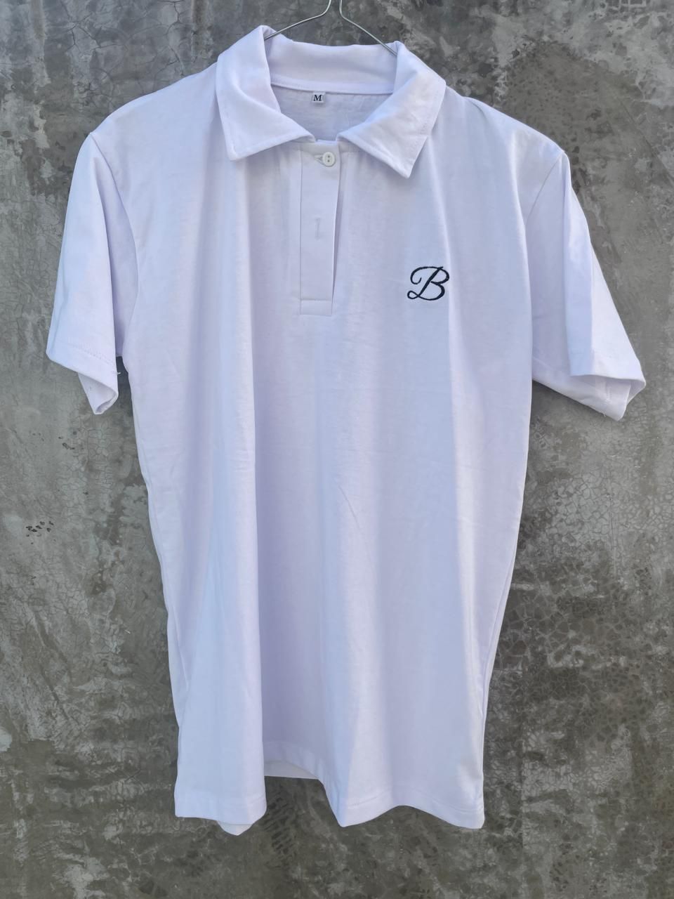 Una camisa polo blanca con la letra b está colgada en una percha.