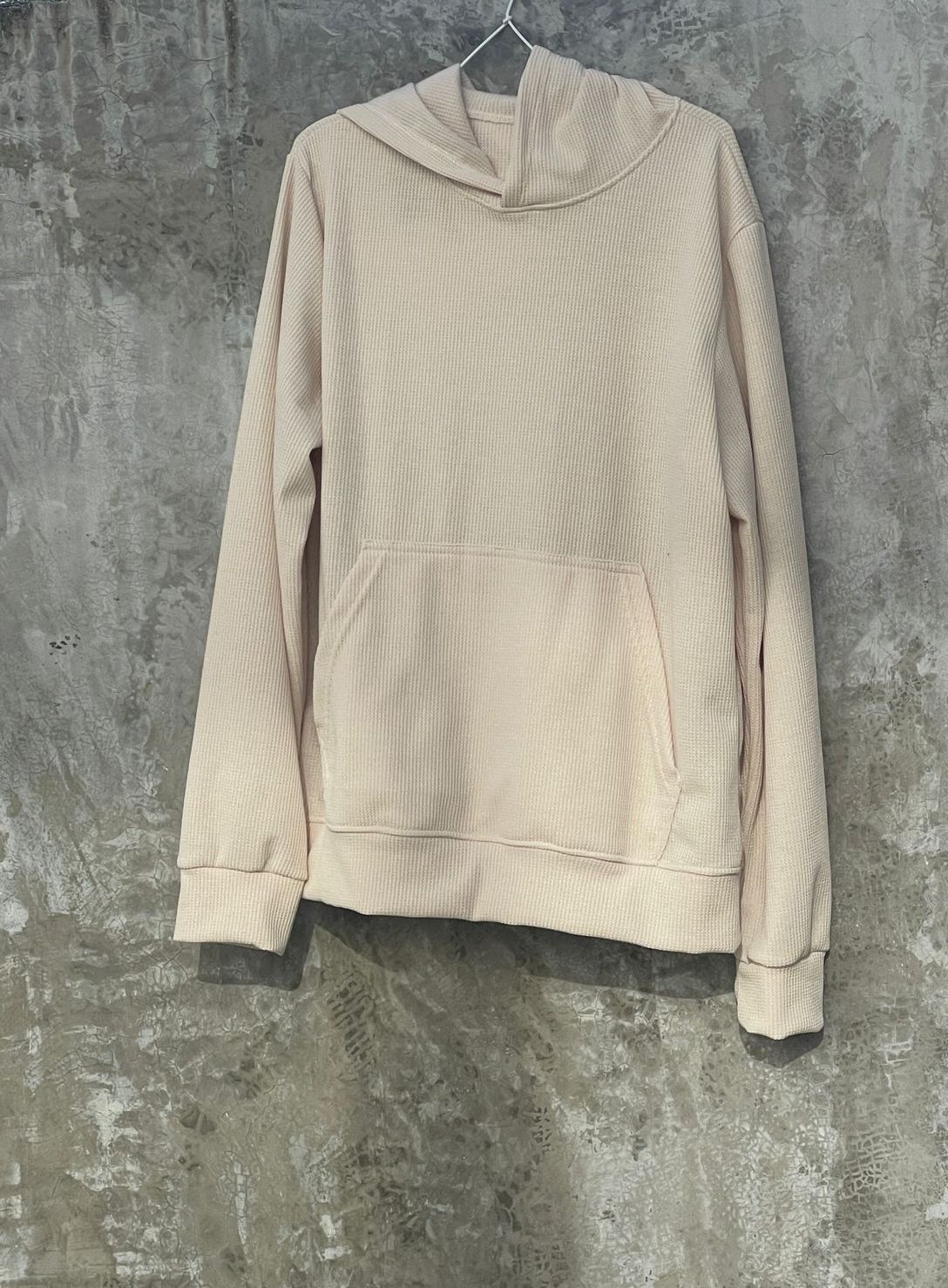 Una sudadera con capucha de color beige cuelga de una percha frente a una pared de hormigón.
