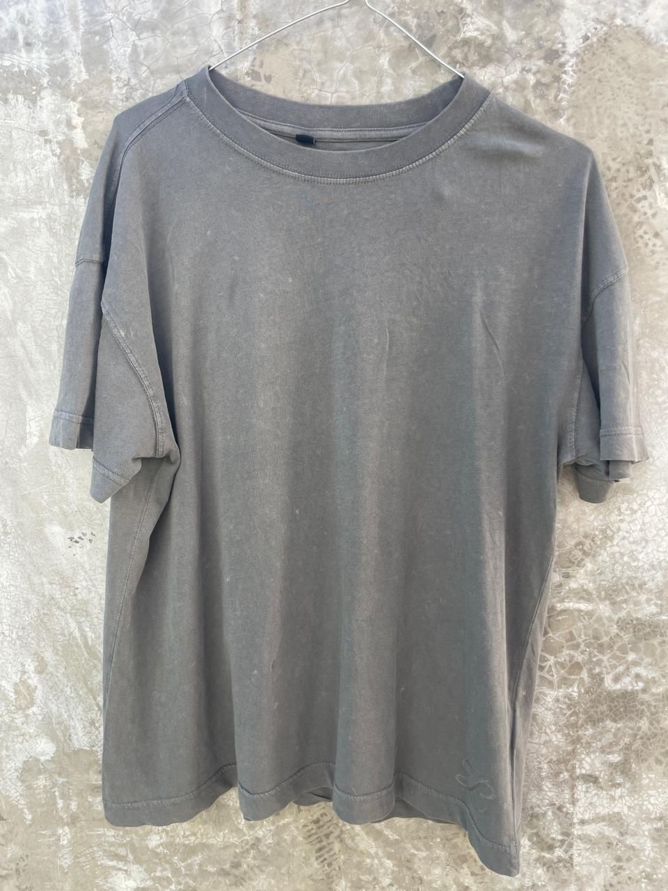 Una camiseta gris está colgada en una percha contra una pared de hormigón.