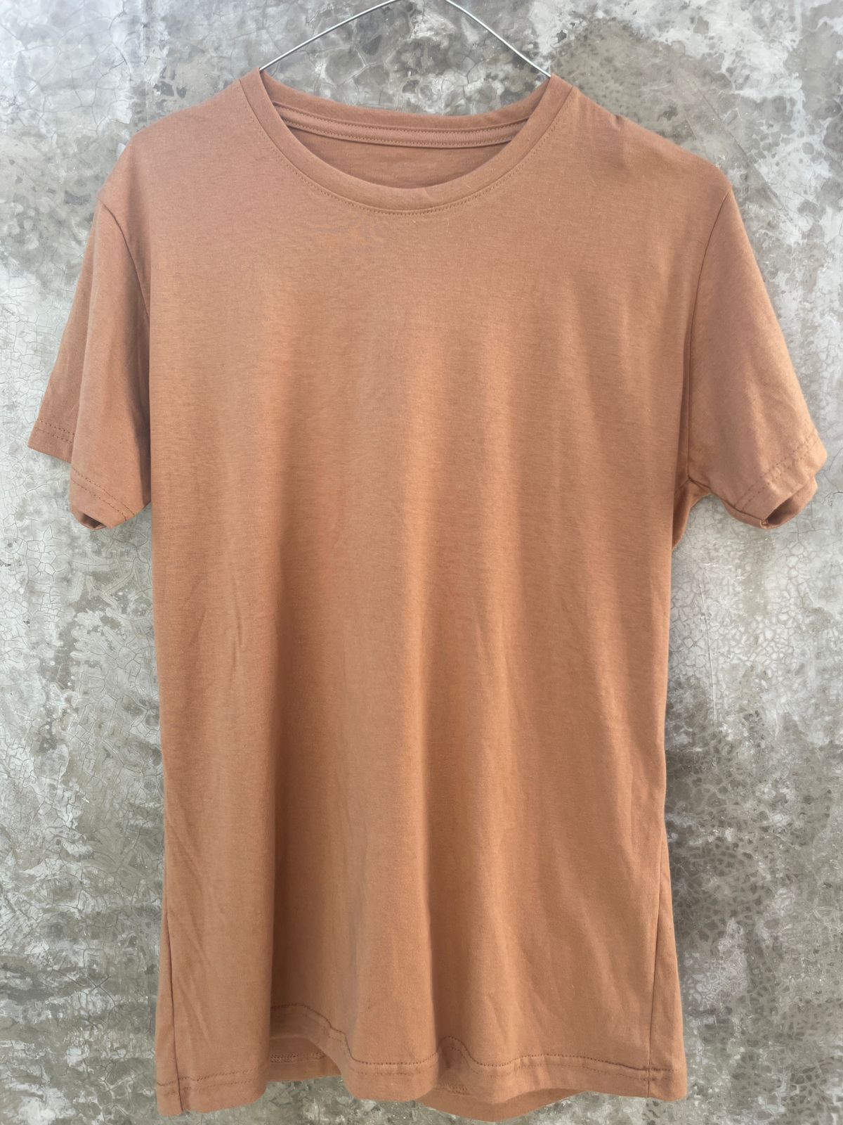 Una camiseta marrón está colgada en una percha contra una pared de hormigón.