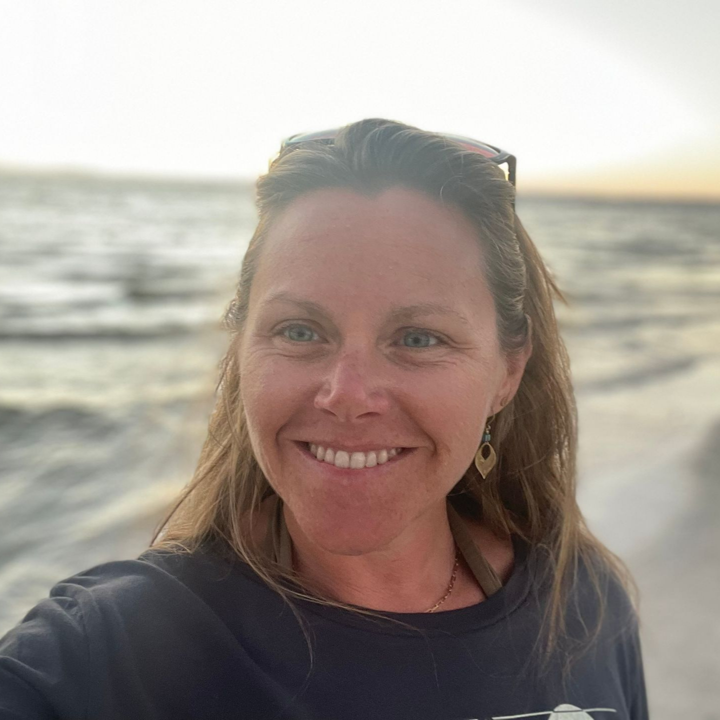 Joni smiling at the camera on a beach; ocean and sunset in the background.