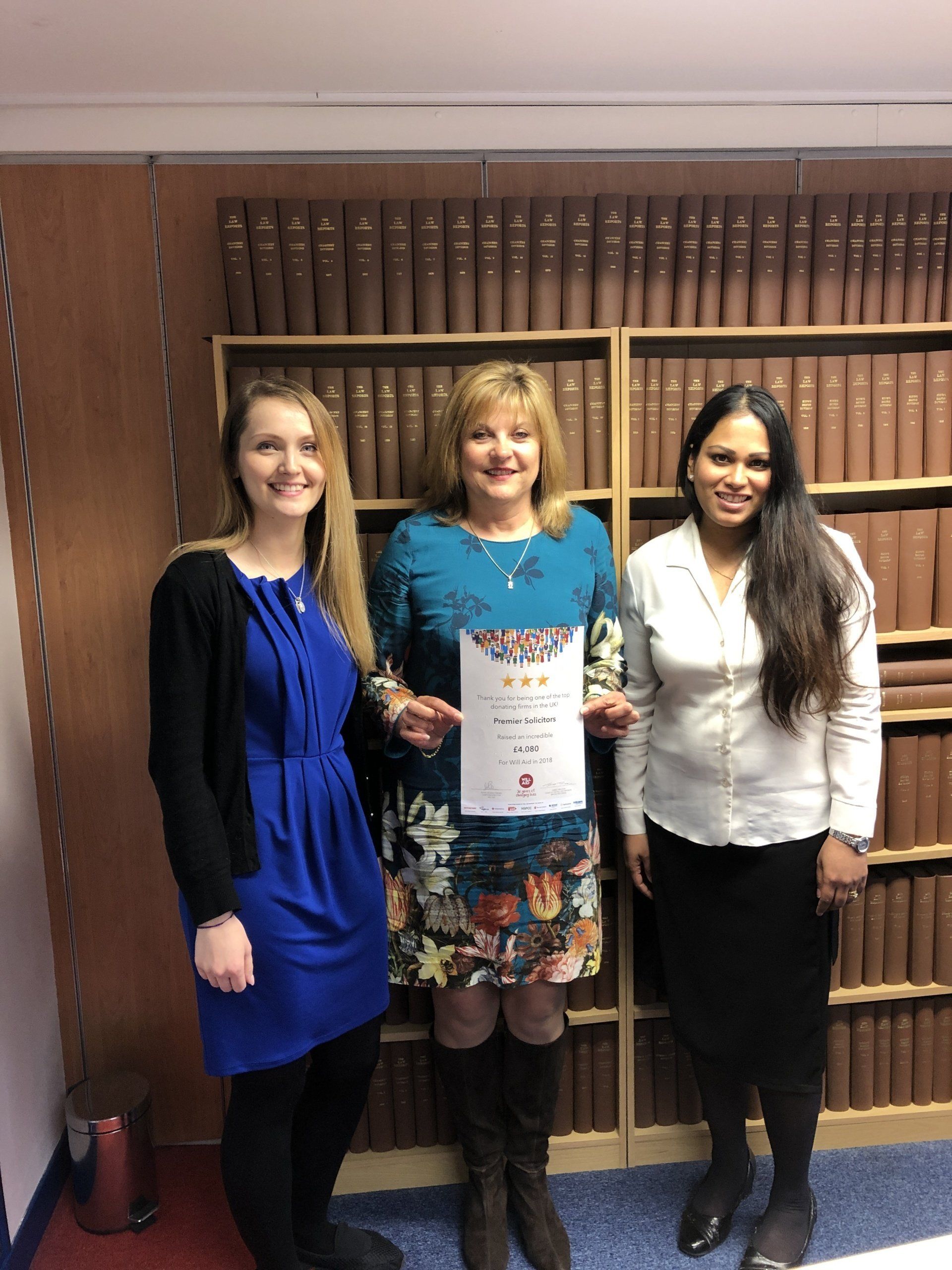 Three women are standing in front of a bookshelf holding a certificate