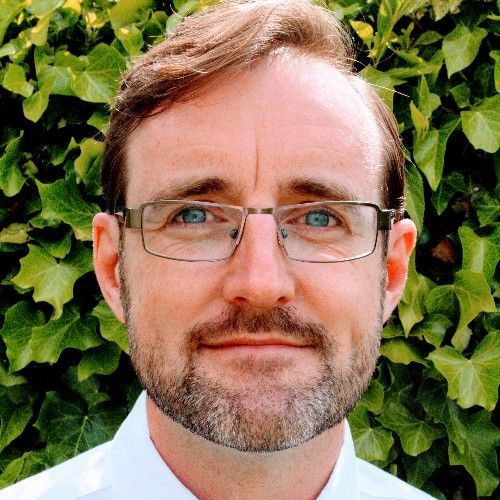 A man with glasses and a beard is standing in front of a bush.