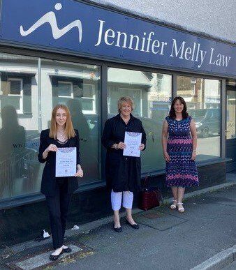 Three women are standing in front of a building holding certificates.