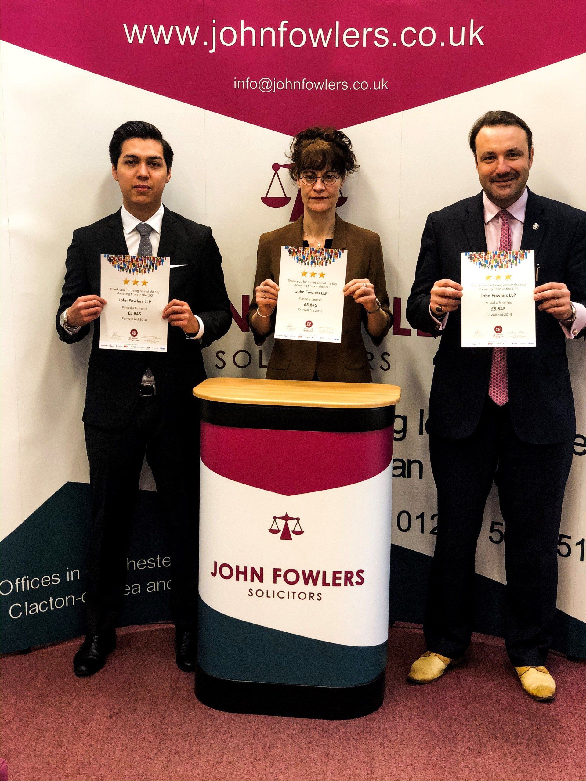 Three people holding papers in front of a john fowlers sign