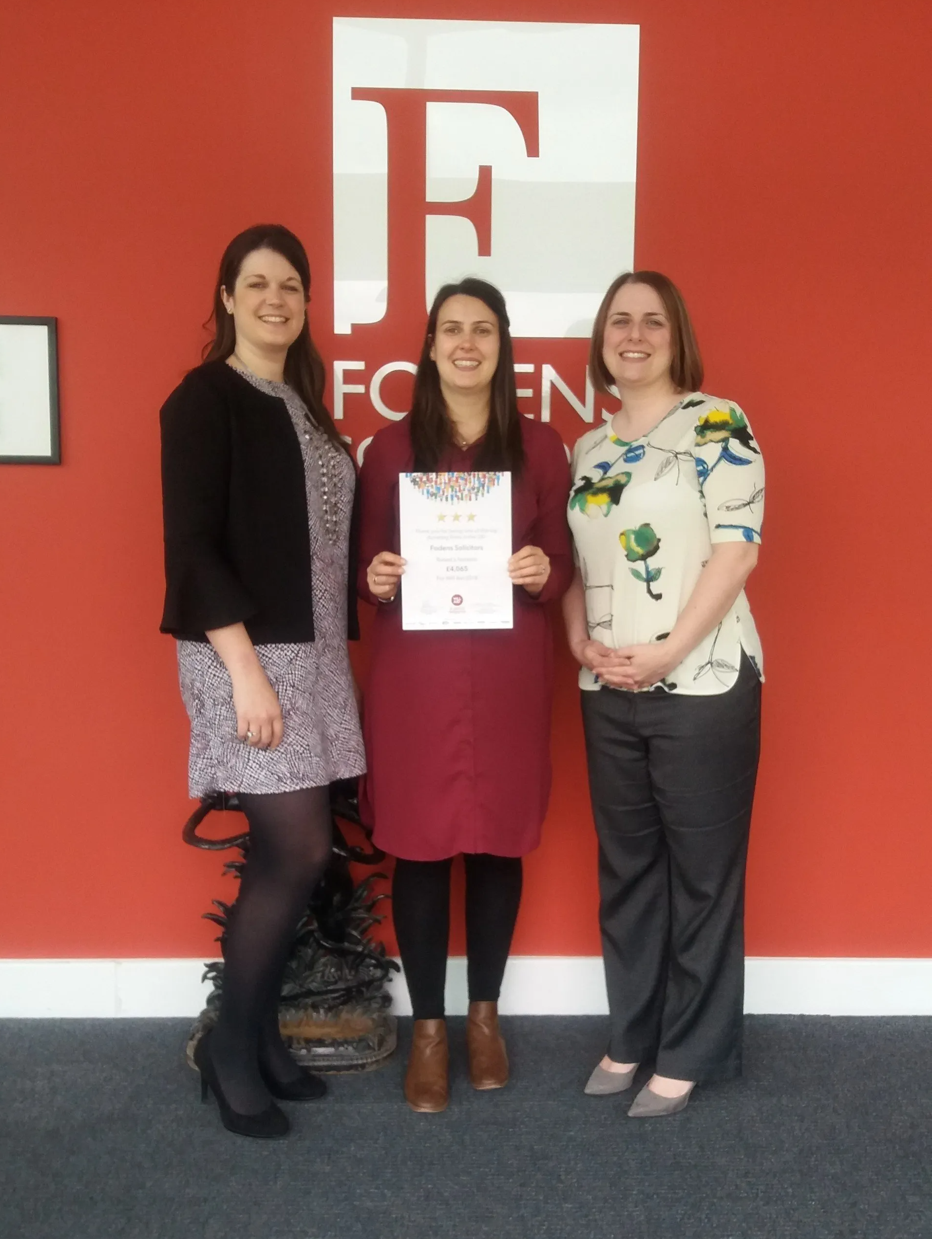 Three women stand in front of a red wall with the letter f on it