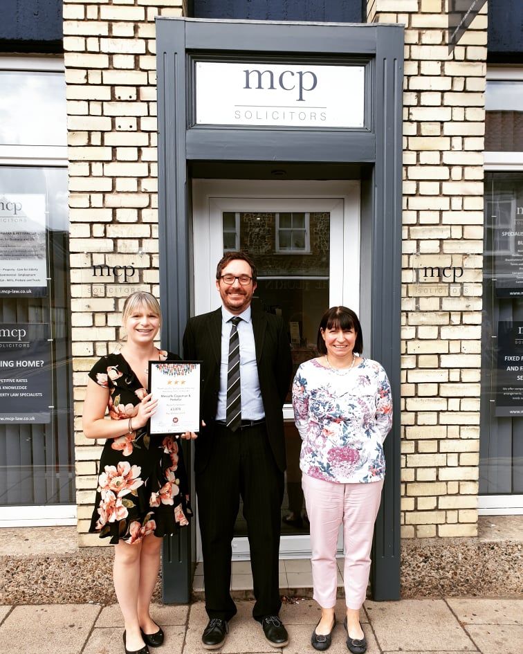 A man and two women are standing in front of a brick building holding a certificate.